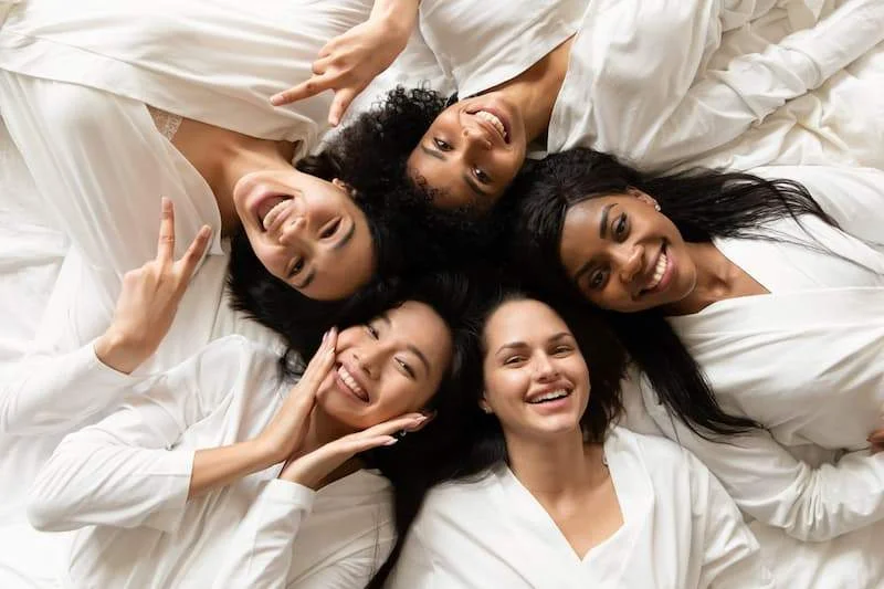 Top view five diverse women in white bathrobes lying in bed smiling and looking at the camera