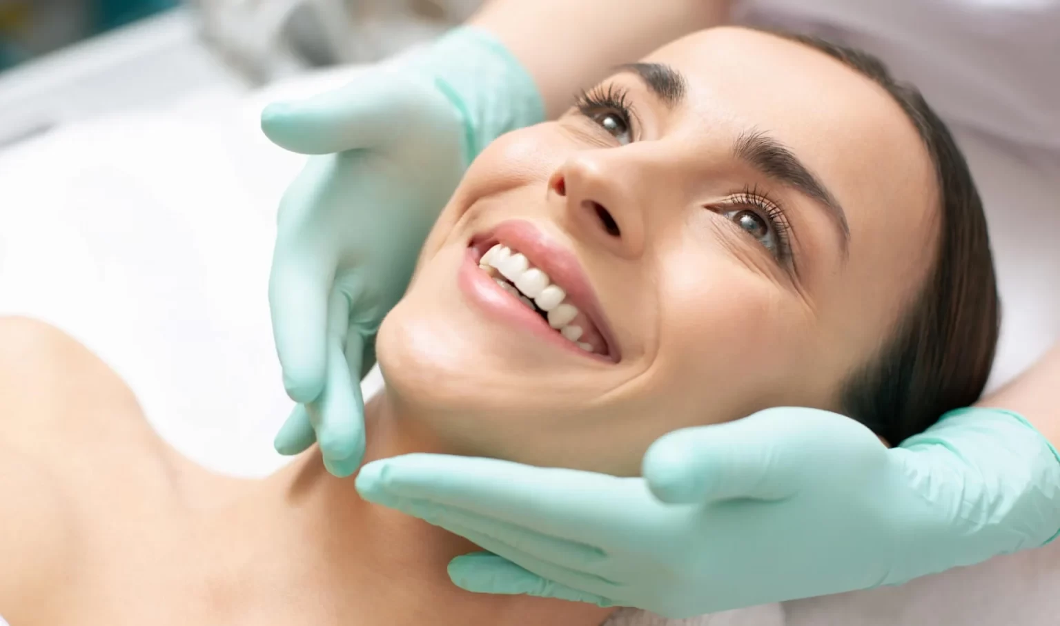 Close up photo of the face of smiling woman and hands in green rubber gloves touching it gently