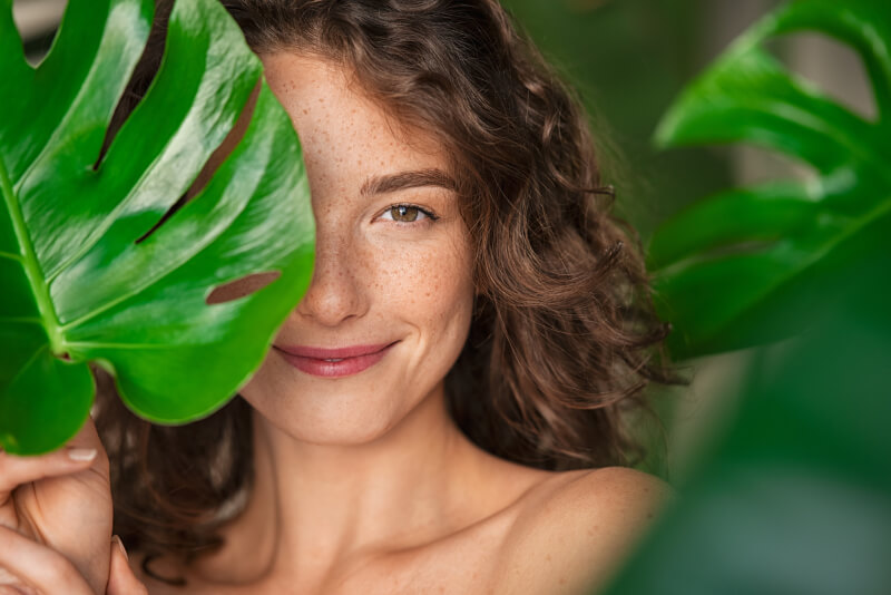 Close up face of beautiful young woman covering her face by green monstera leaf while looking at camera