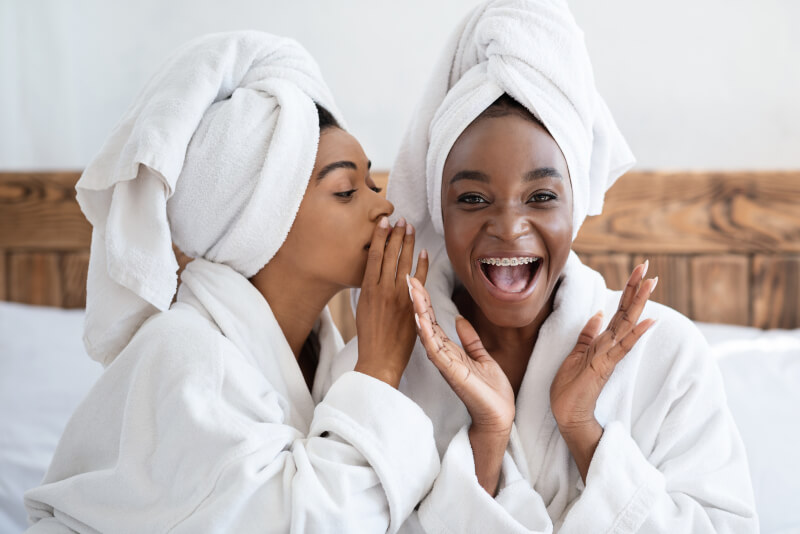 Two pretty african american young women having girls day at home, wearing bathrobes, sitting on bed
