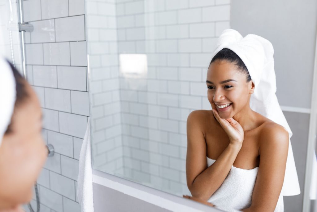 Beautiful young woman looking at her reflection in the mirror, in towels wrapped around head and body