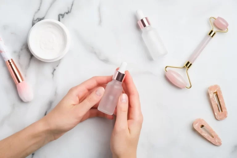 Female hand holding serum bottle over marble table with moisturizer cream makeup brush face massage roller