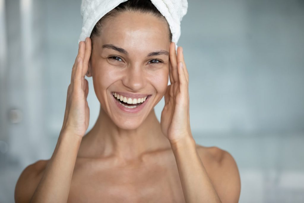 Head shot of happy woman with head wrapped in bath towel, touching face with toothy smile