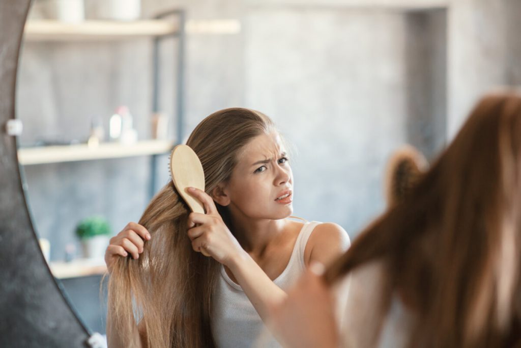 Frustrated young woman trying to brush her tangled hair in front of mirror at bathroom