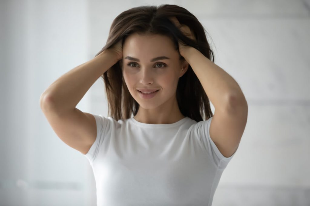 Long-haired millennial woman standing by mirror burying hands to her lovely shiny hair