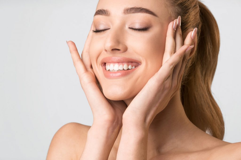 Cheerful Blonde Girl Touching Face Posing On Gray Studio Background