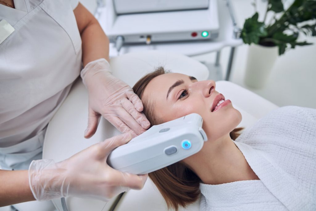 Close up side view portrait of young female lying on medical chair while unrecognized provider making non surgical face lifting in medical spa