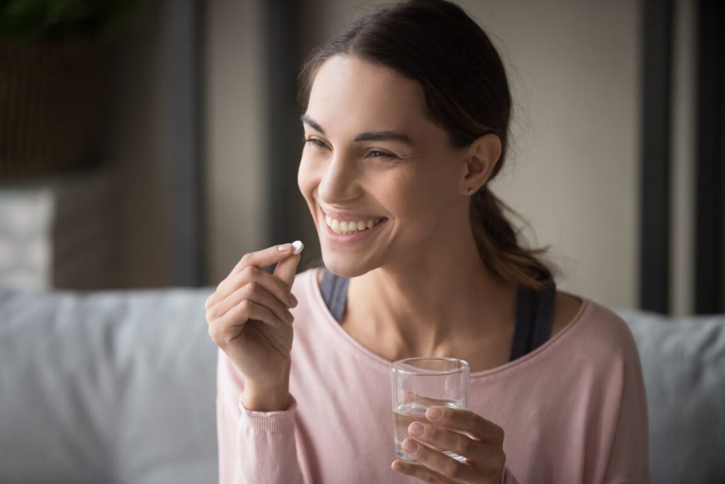 woman holding pill for hait loss with glass of water while sitting on sofa at home