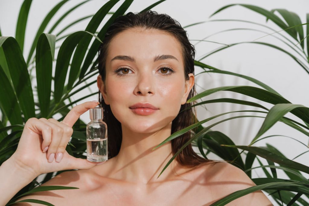 Beauty portrait of a young white woman standing over green leaves showing bottle of facial oil