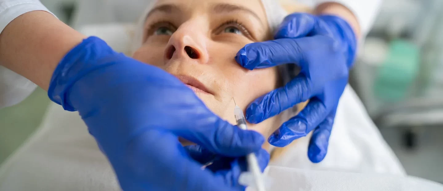 A female client lying down and wearing a protective gown is receiving a botox injection from a pair of hands wearing blue surgical gloves.