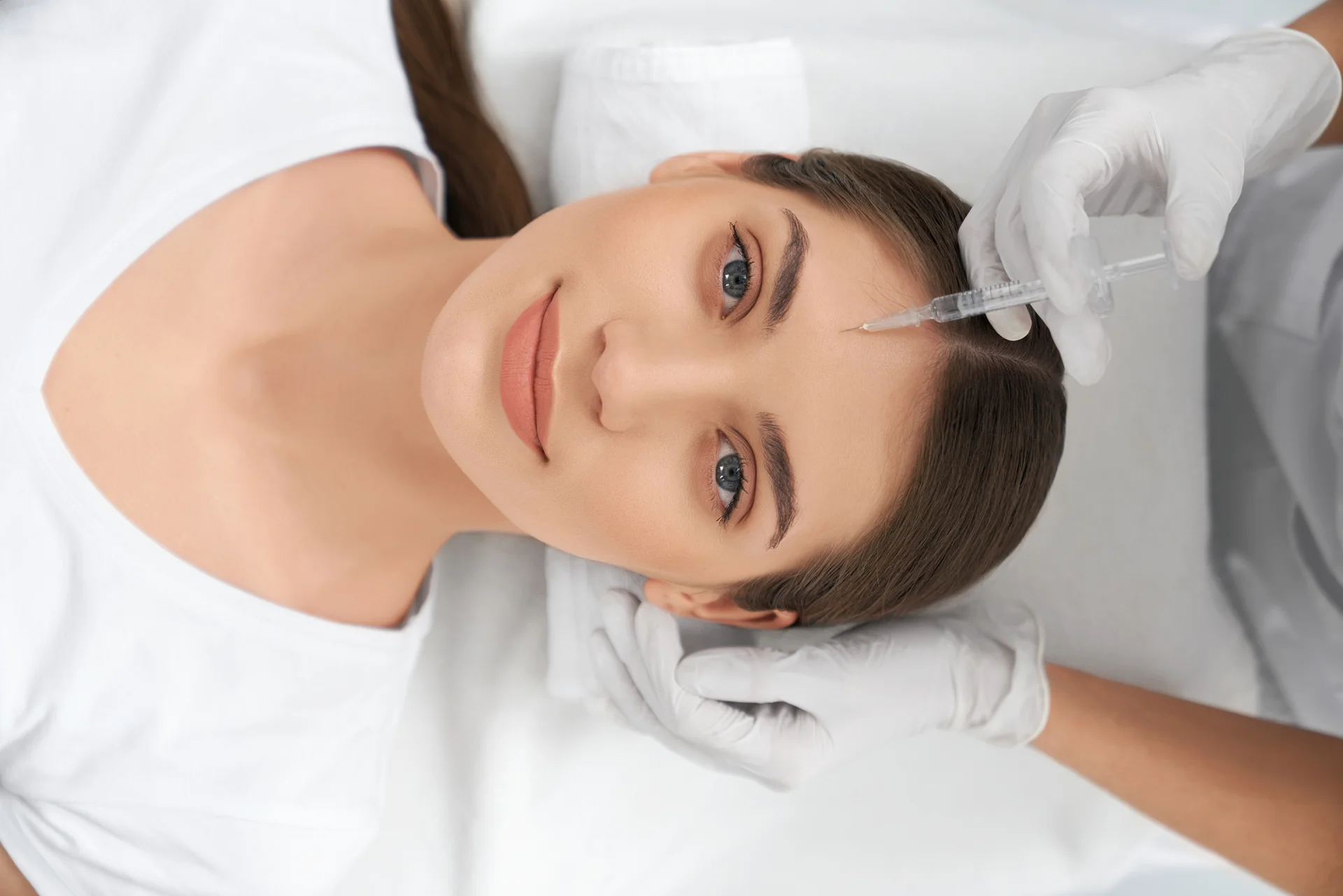 Top view portrait of cute young woman lying on procedure for face skin improvements thru injection anti-aging skin with professional cosmetics.
