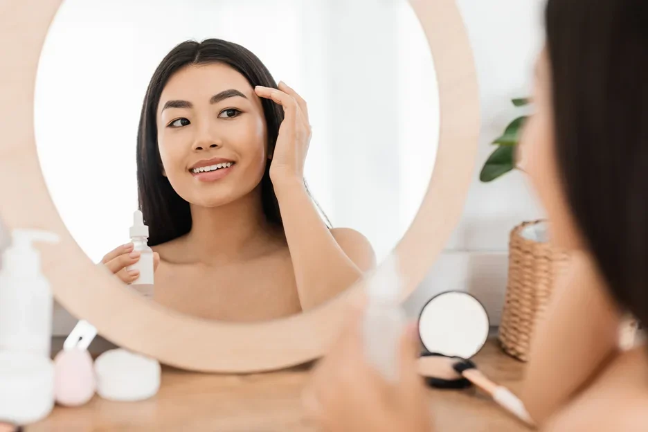 a woman checks her face in front of the mirror while holding a bottle of pdrn caffeine shot serum