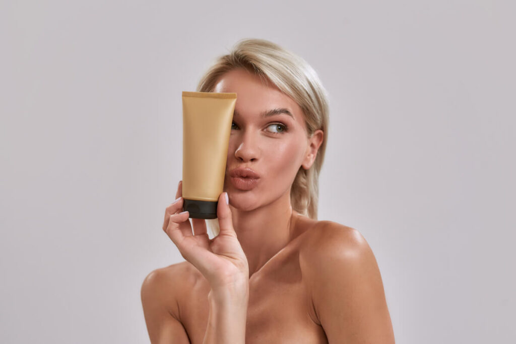 Portrait of cheerful young woman with perfect glowing skin looking aside, holding sunscreen skincare product
